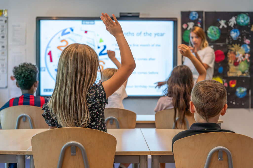 Unga skolbarn sitter vid sina skolbänkar i ett klassrum, en flicka räcker upp handen